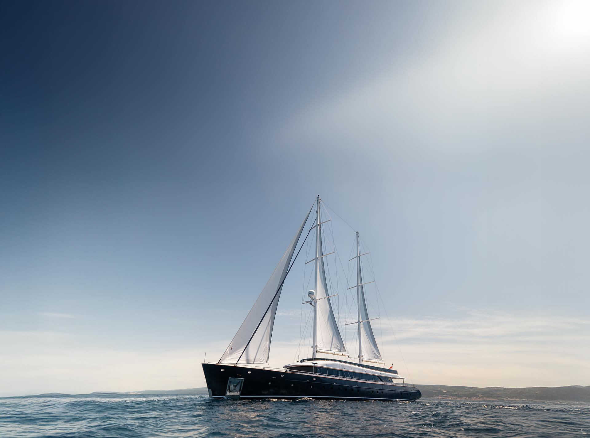 Luxury black sailing yacht with two tall masts and white sails gliding across open water beneath a wide, clear blue sky.