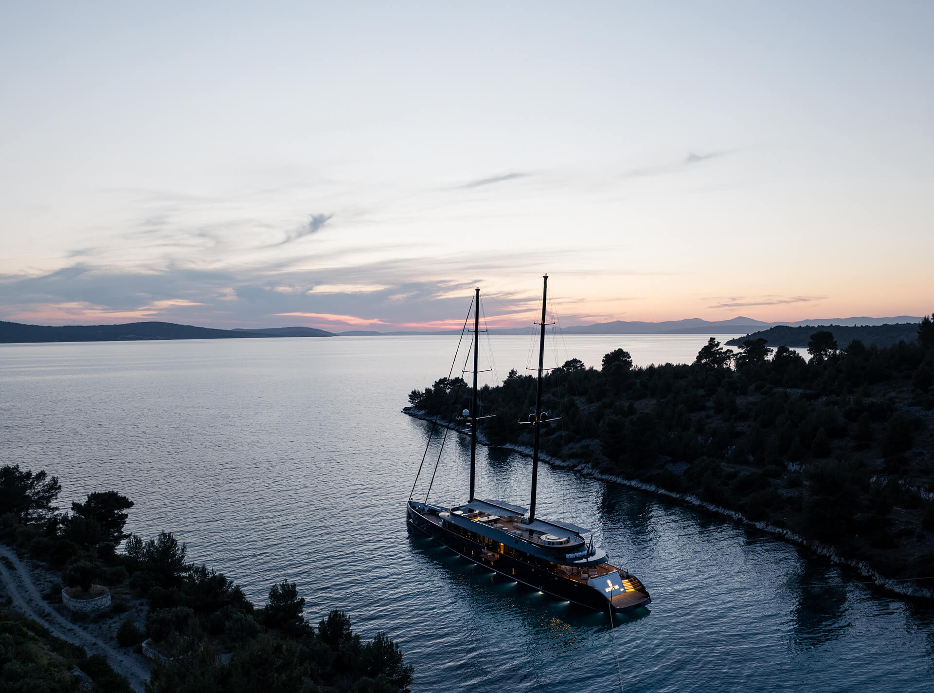 Aerial view of a luxury sailing yacht anchored in a quiet bay at sunset, surrounded by forested coastline and calm water with soft evening light on the horizon.