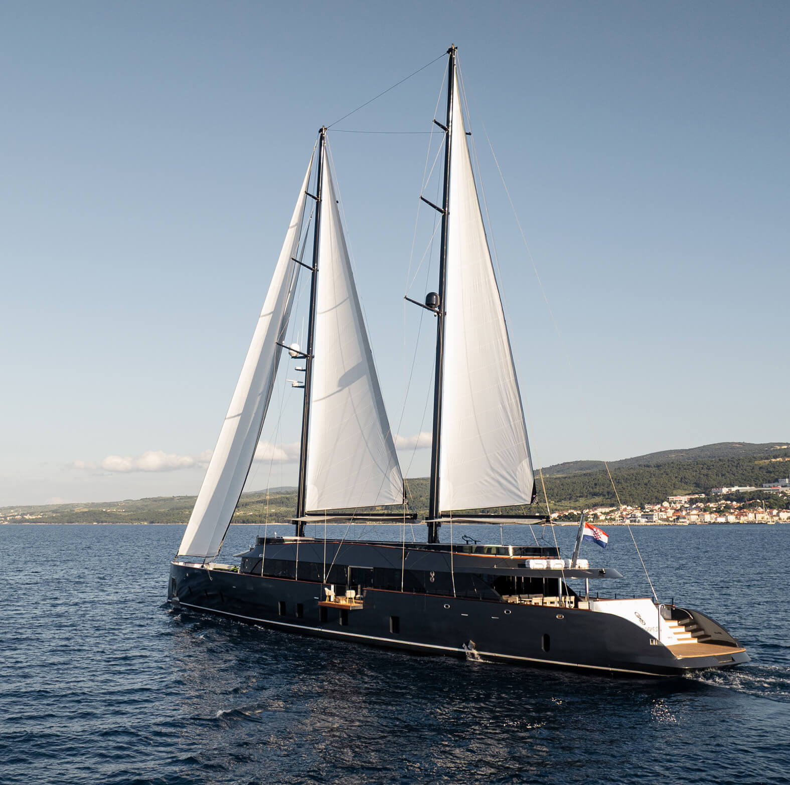 Large modern sailing yacht with two tall masts and white sails cruising on calm sea near a coastal town and green hills under a clear sky.