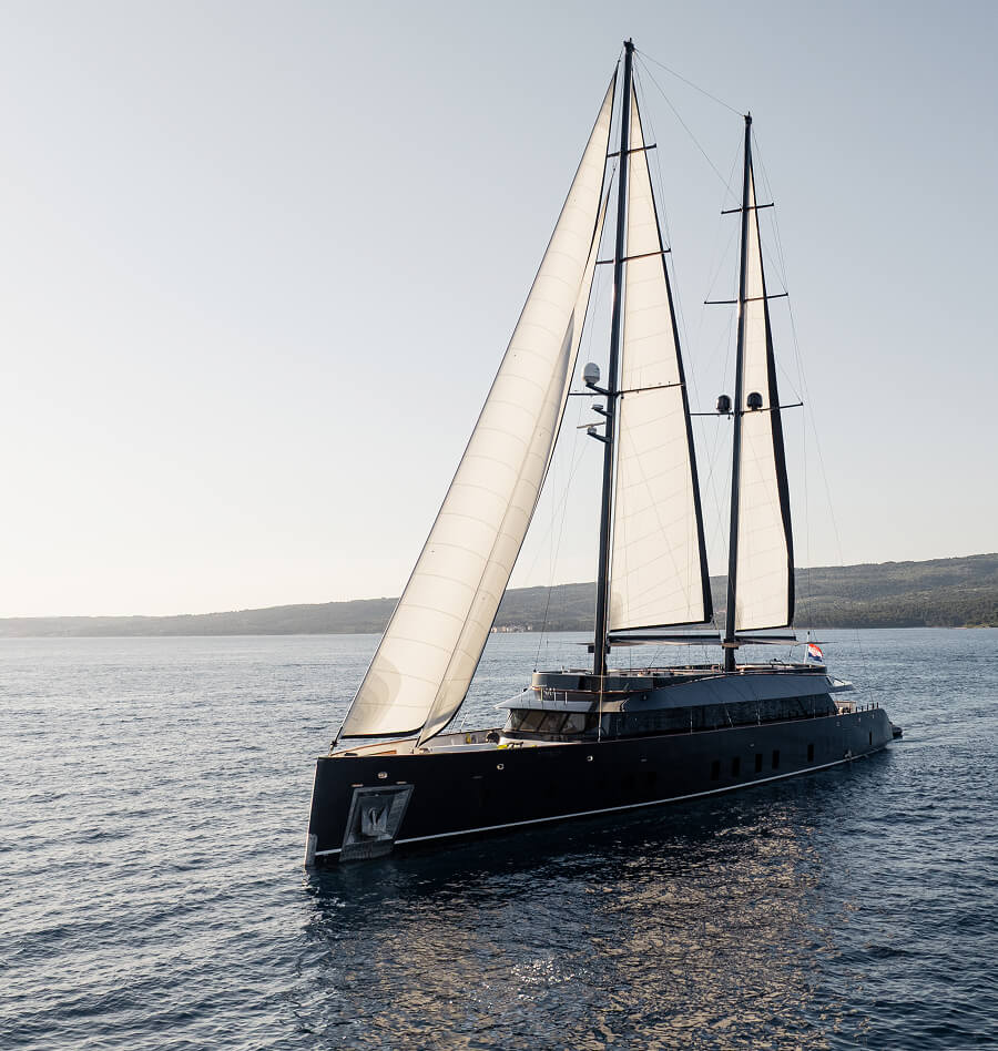 Modern black sailing yacht with two tall masts and white sails gliding on calm sea near a low, green coastline under a clear sky.