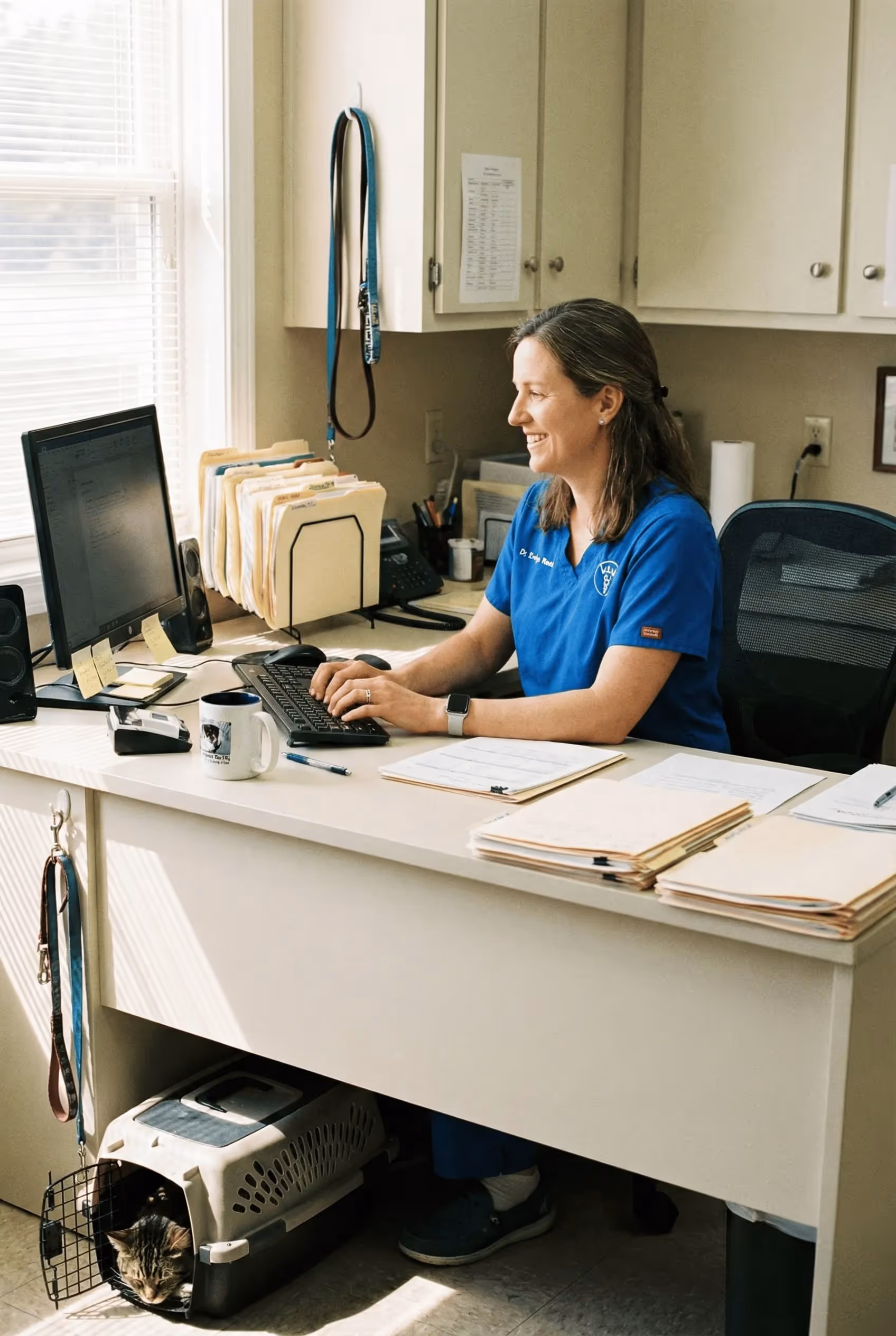 A female veterinarian in blue scrubs types on a keyboard at her desk while a cat rests in a pet carrier underneath.