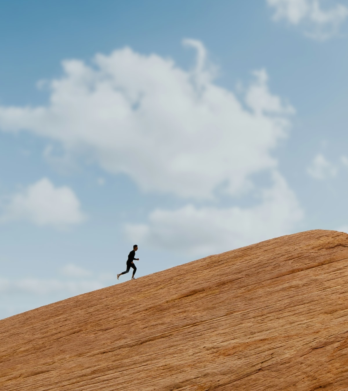 Person running uphill on a large reddish rock formation under a partly cloudy blue sky.