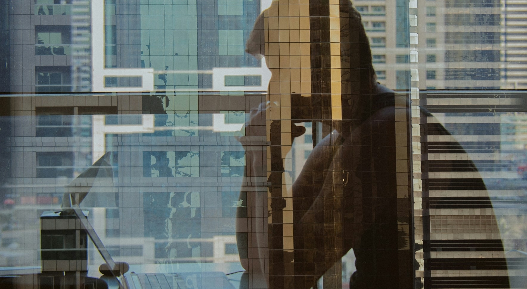 Silhouette of a person sitting at a desk with a laptop, overlaid with reflections of tall city buildings.