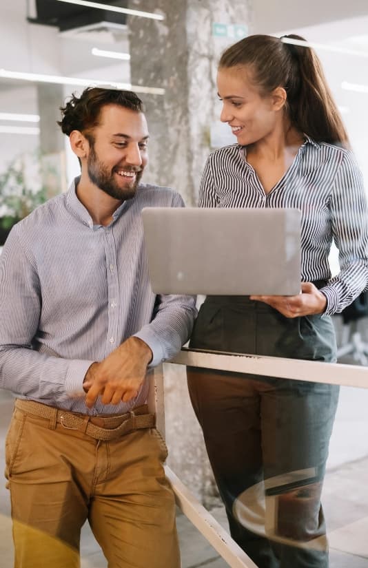 Man and woman in business casual attire smiling and looking at a laptop in an office setting.