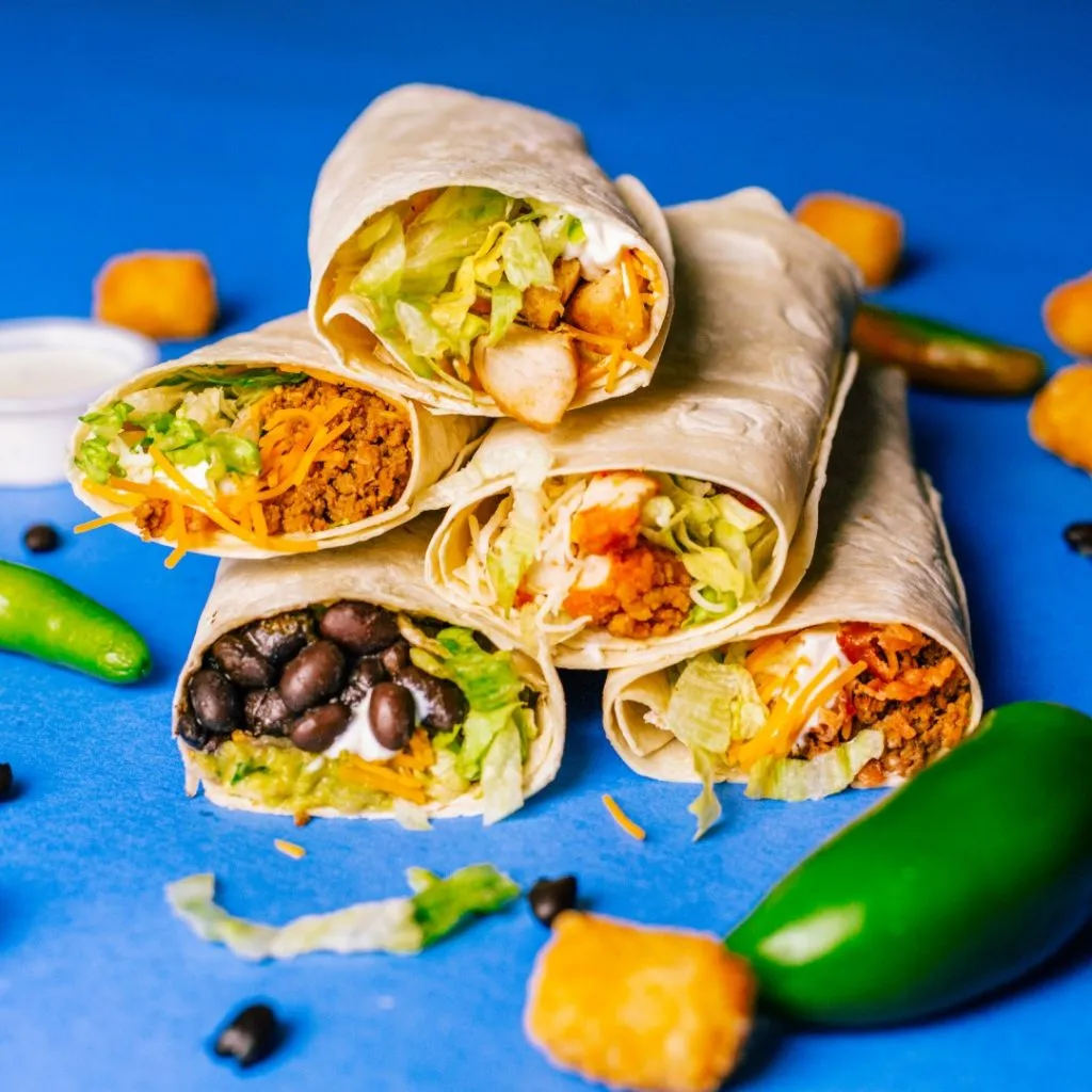 A stack of five burritos filled with ingredients like black beans, lettuce, and cheese on a blue background, surrounded by jalapeños and sauce.