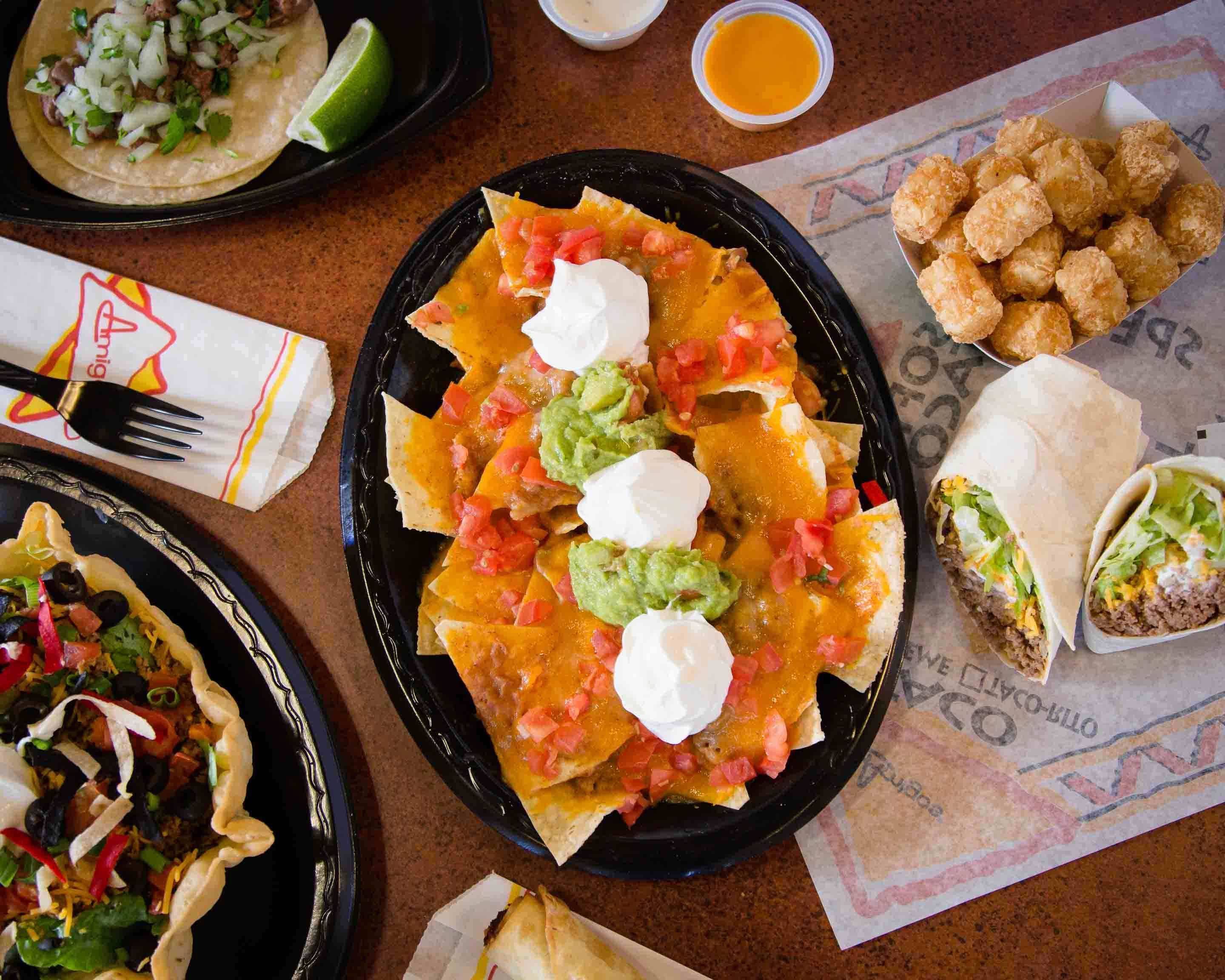 A vibrant spread of Amigos food includes nachos topped with cheese, guacamole, and sour cream, a burrito, crispy tots, and a taco salad on a colorful table.