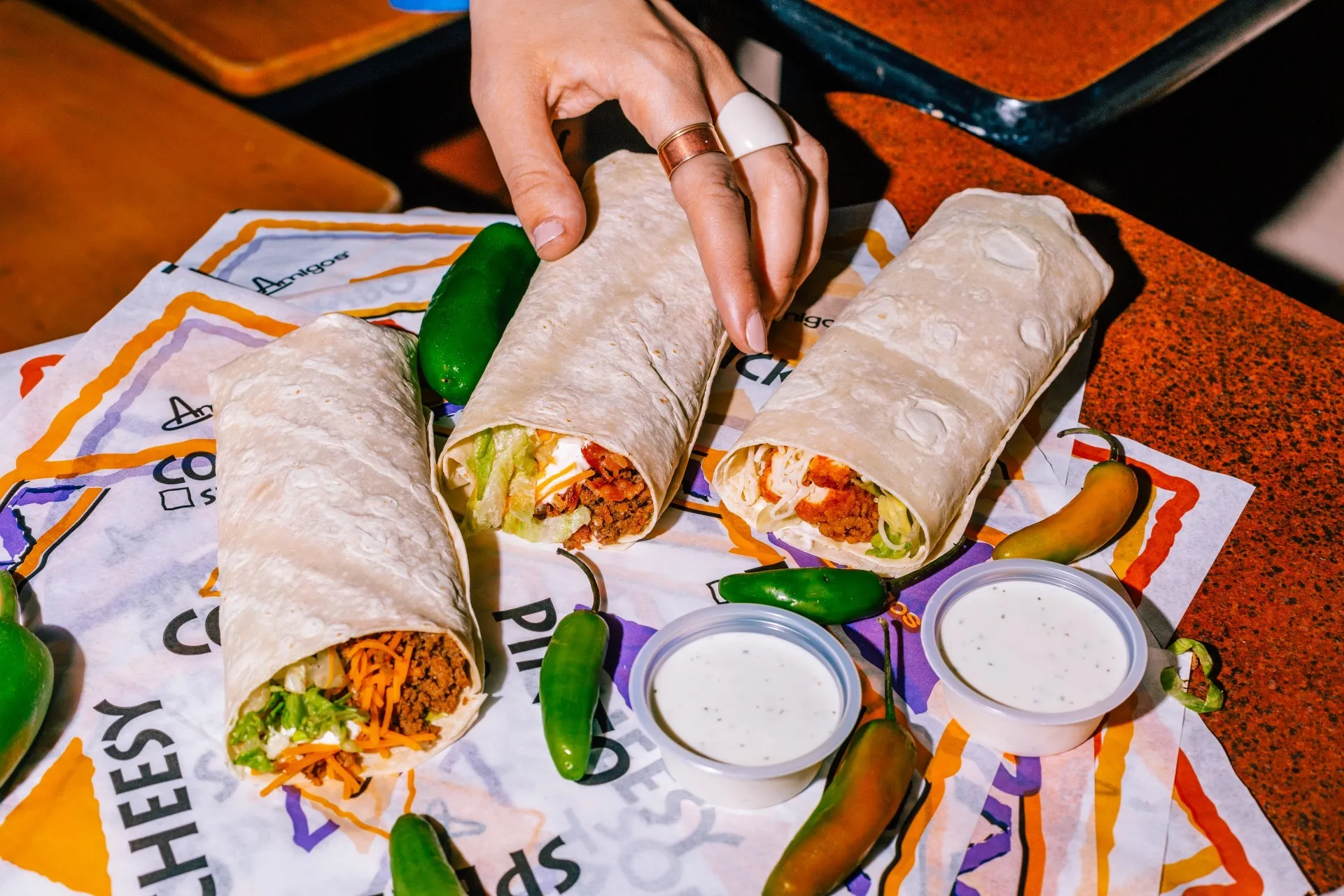 A hand reaches for one of three burritos on colorful wrapper paper, surrounded by green chilies and two cups of white dipping sauce, conveying a vibrant, appetizing scene.