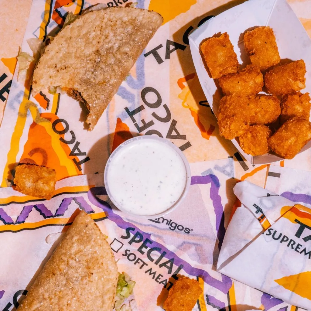 Taco and tater tots on colorful branded wrappers with a cup of ranch dressing in the center. Casual and vibrant fast-food presentation.