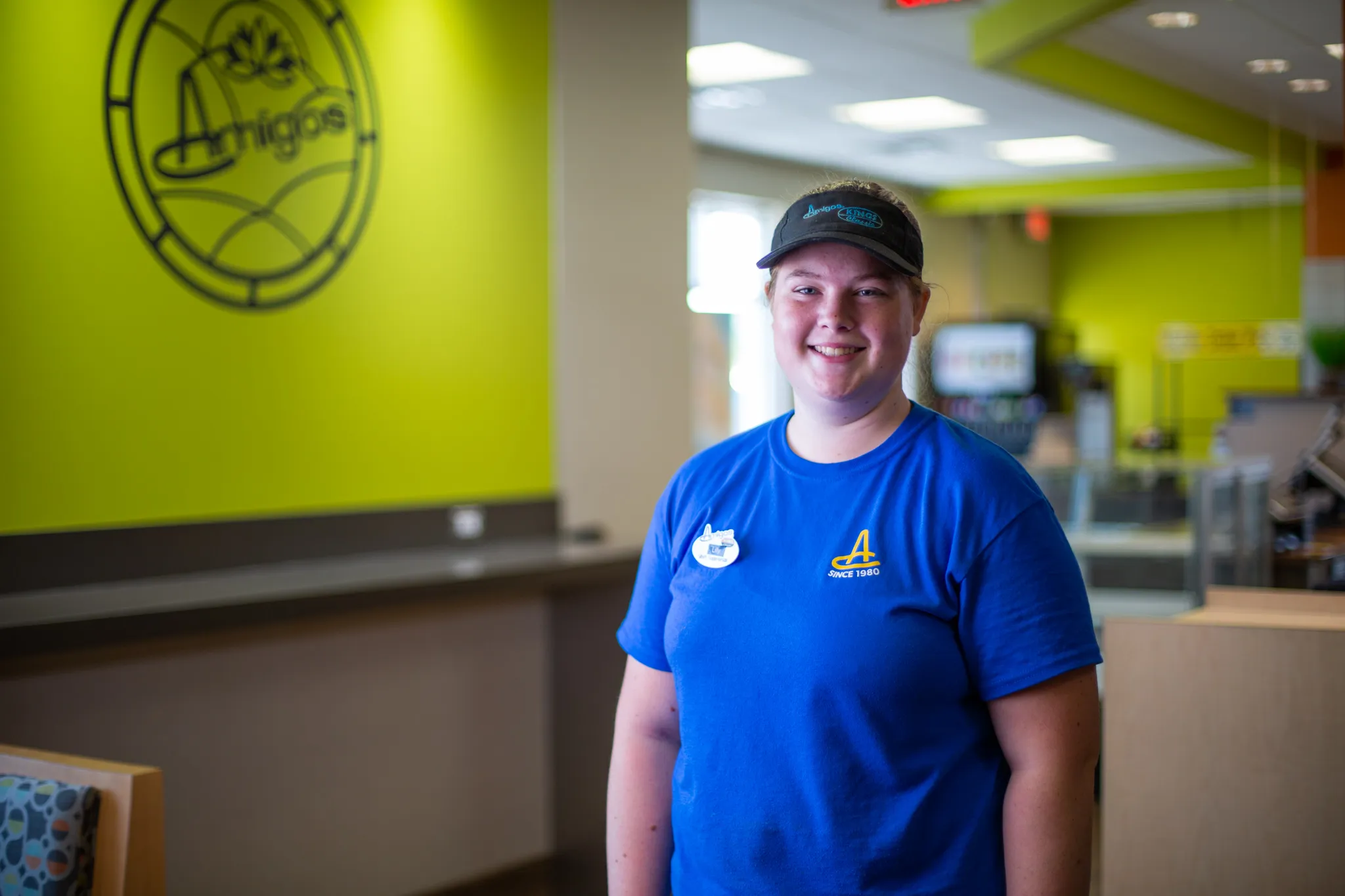 Smiling person in a blue shirt and cap stands inside a brightly lit café with lime green walls and modern decor, creating a welcoming atmosphere.