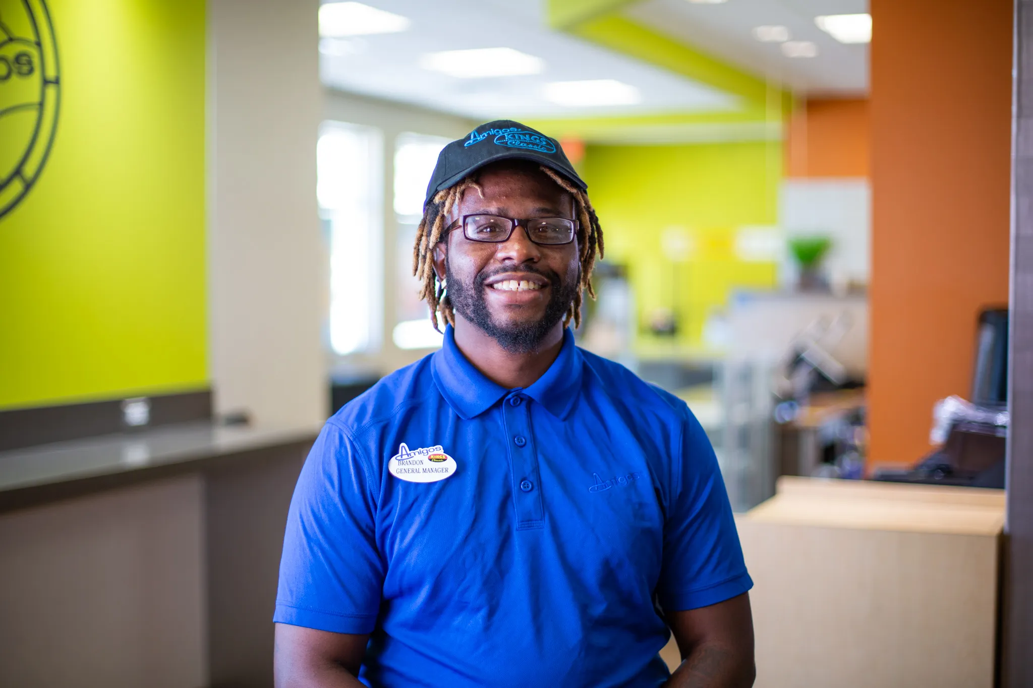A smiling person wearing a blue polo and cap stands in a brightly lit, colorful office. Their name tag is visible. The atmosphere is welcoming.