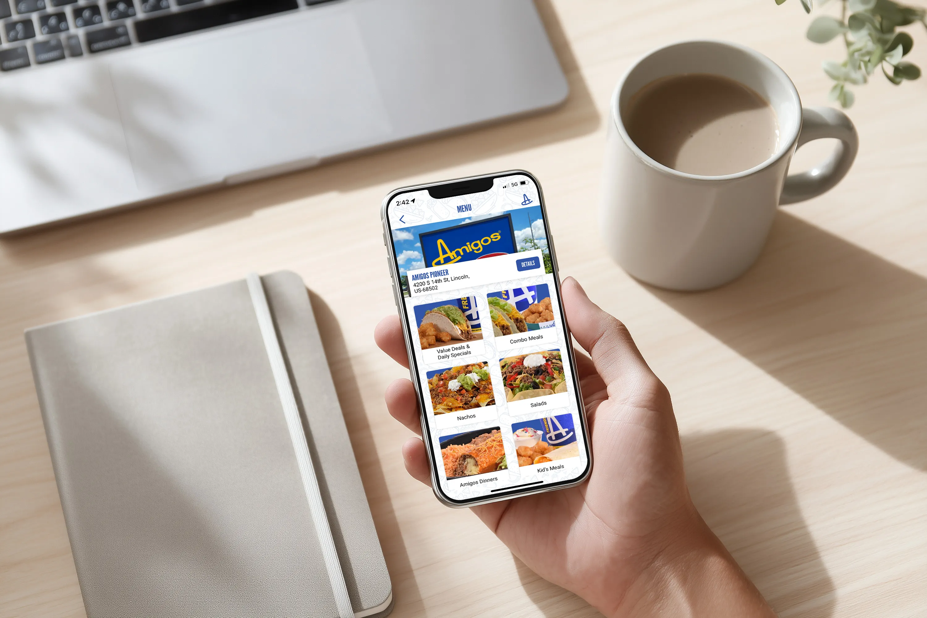 A person holds a smartphone displaying a food delivery app with various dishes. A notebook, open laptop, and coffee cup are on the bright wooden table.
