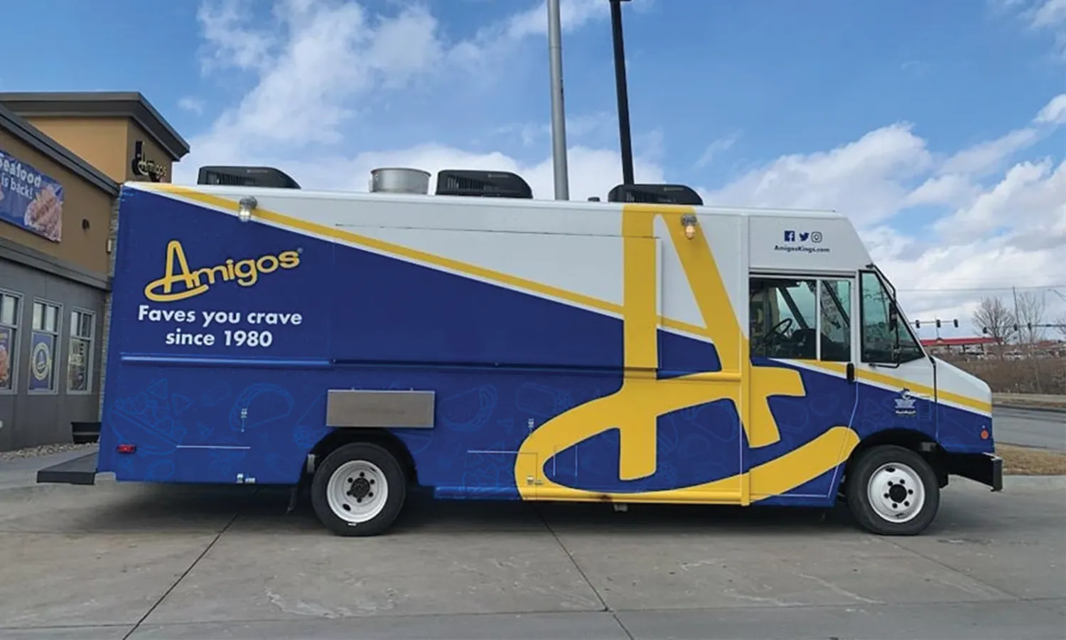 A vibrant blue and white food truck with bold yellow accents is parked on concrete. The truck displays "Amigos" and "Faves you crave since 1980" on the side. A clear, sunny sky is in the background.