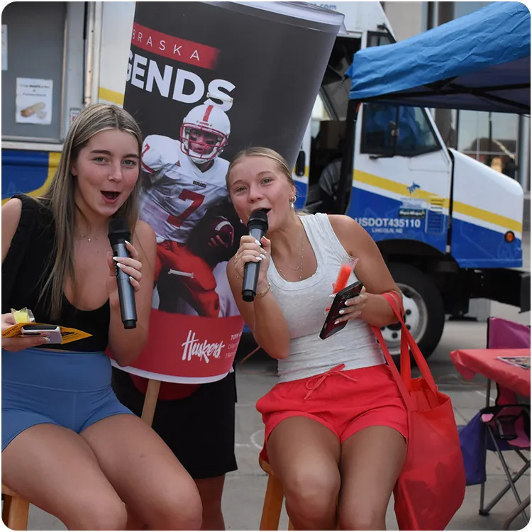 Two young women sitting on stools holding microphones and smiling, with a person in a Nebraska Legends football cup costume behind them.