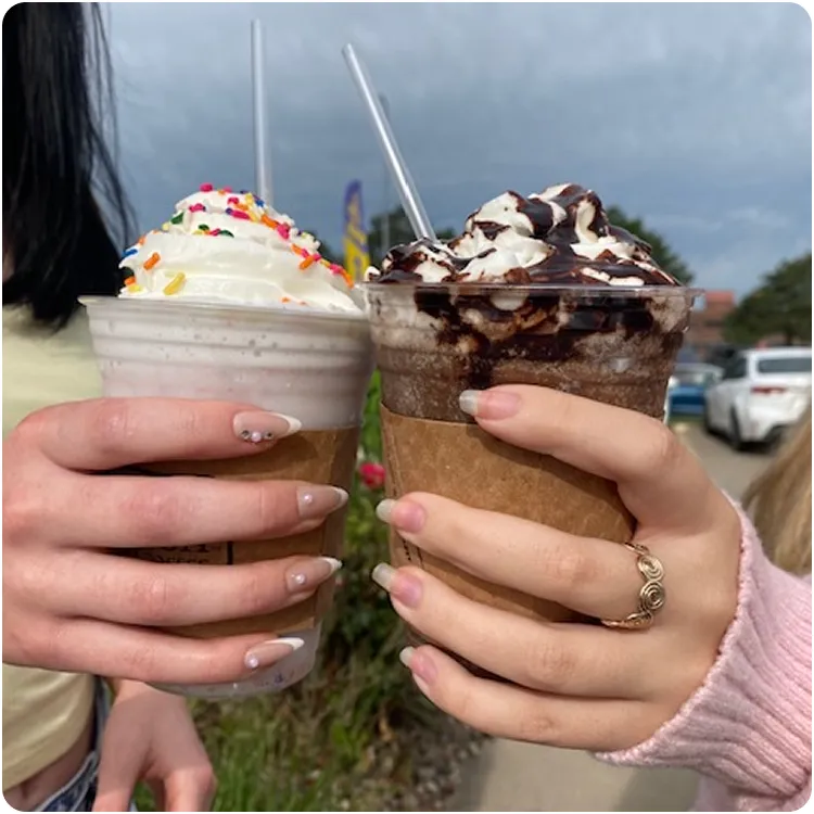 Two hands holding iced blended coffee drinks topped with whipped cream; one with colorful sprinkles and the other with chocolate syrup.