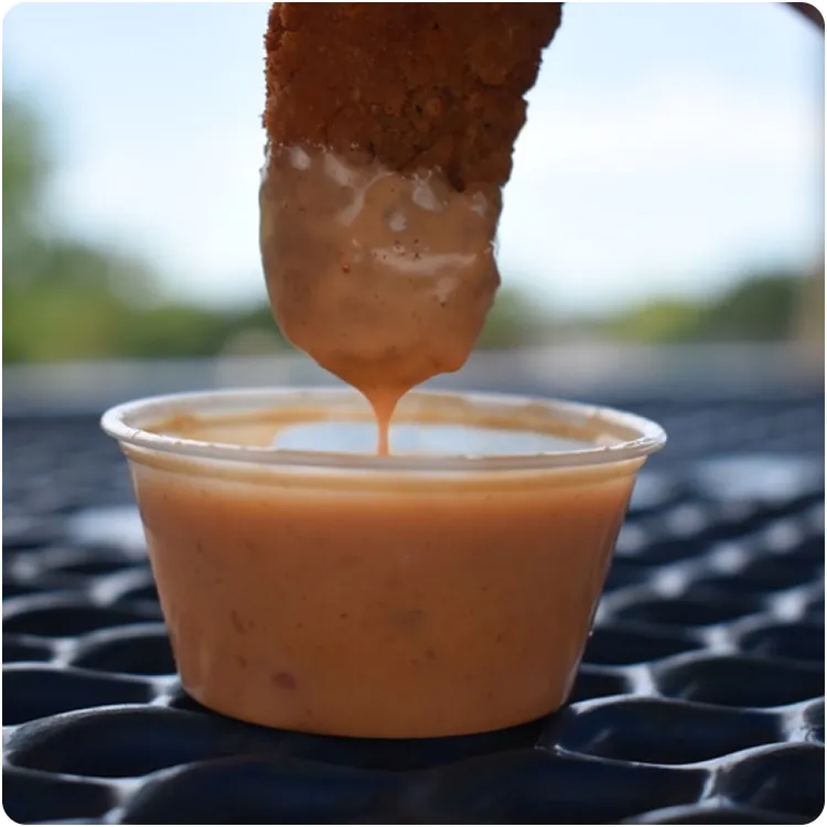 Crispy fried food piece being dipped into a small cup of creamy orange sauce on a textured blue surface.