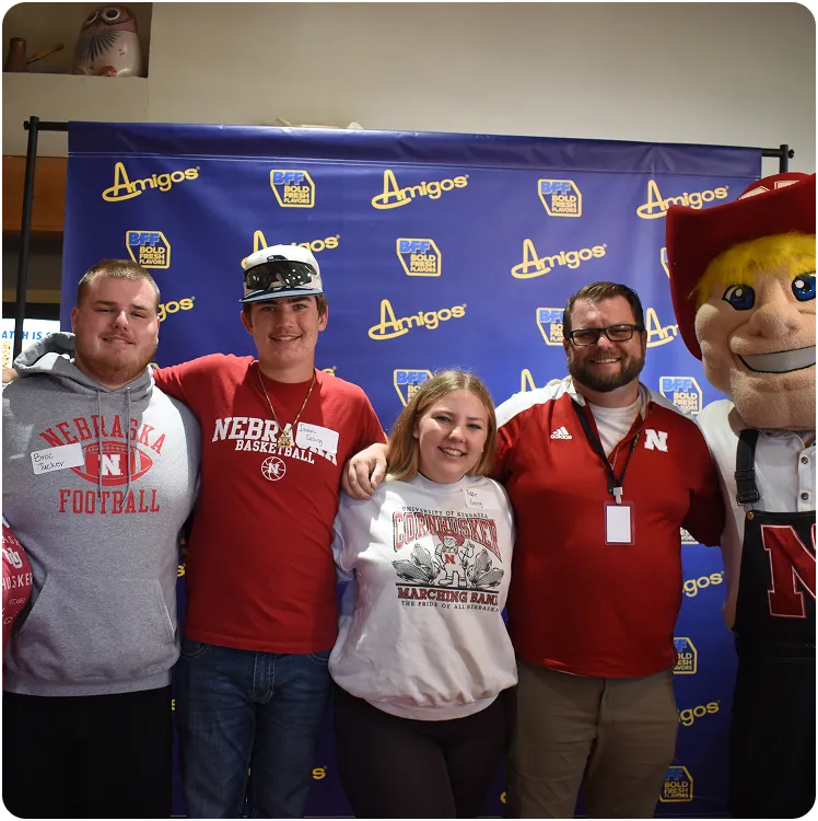 Four people smiling in front of a blue Amigos banner, including individuals wearing Nebraska sports apparel and a mascot dressed in red and white with a large smiling face.