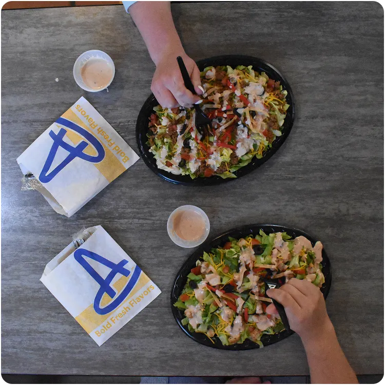 Two hands holding forks and eating taco salad bowls with lettuce, tomatoes, olives, cheese, and dressing on a gray table with two folded taco shell wrappers and two dipping sauces.