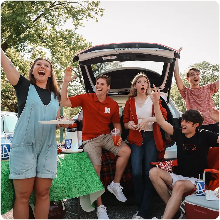 Five happy young adults enjoying a tailgate party around an open car trunk with drinks and snacks.