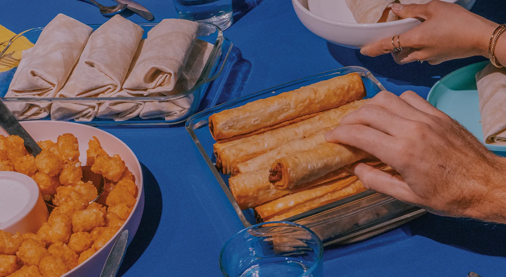 Two hands reaching for taquitos in a glass dish on a blue tablecloth, surrounded by rolled tortillas and a bowl of tater tots with dipping sauce.