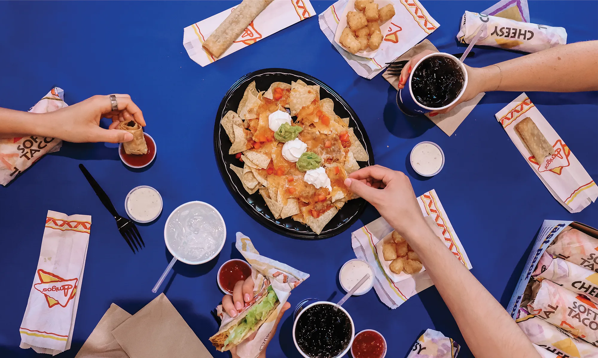 Overhead view of a table with nachos topped with cheese, sour cream, and guacamole, surrounded by tacos, drinks, dipping sauces, and tater tots, with hands reaching for the food.