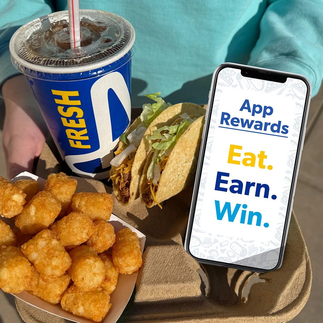 Tray with tater tots, two tacos with lettuce and cheese, a blue cup with soda and straw, and a smartphone displaying 'App Rewards Eat. Earn. Win.'