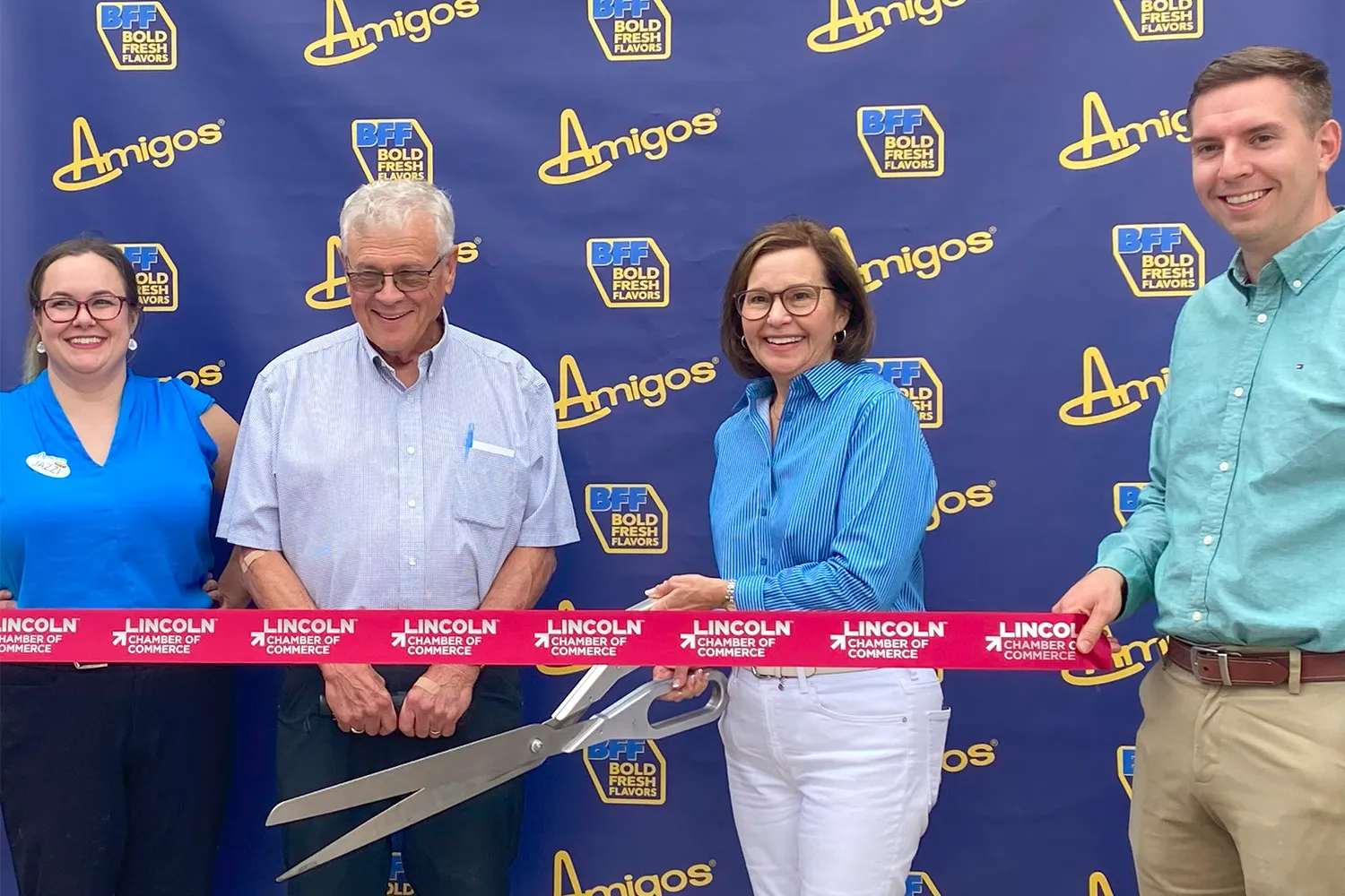 Four smiling people standing in front of an Amigos and BFF Bold Radio backdrop cutting a red Lincoln Pharmacy ribbon with large scissors.