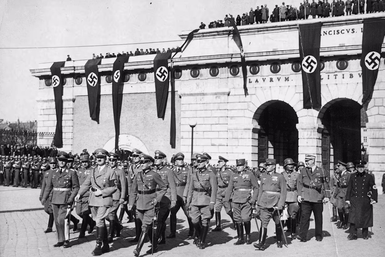 Black-and-white historical photograph showing Adolf Hitler and a group of high-ranking Nazi officials marching in formation in front of a large building draped with multiple Nazi swastika flags. Soldiers stand in formation on either side, and civilians are visible on the rooftop and in the background watching the parade. The scene appears formal and militaristic, emphasizing the display of Nazi power.
