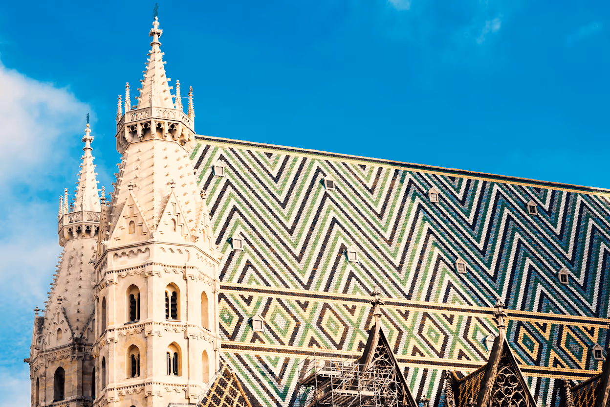 A close-up of St. Stephen's Cathedral in Vienna, highlighting the massive, steep roof with its distinctive chevron pattern of green, black, and white ceramic tiles, next to a Gothic stone spire, set against a bright blue sky.
