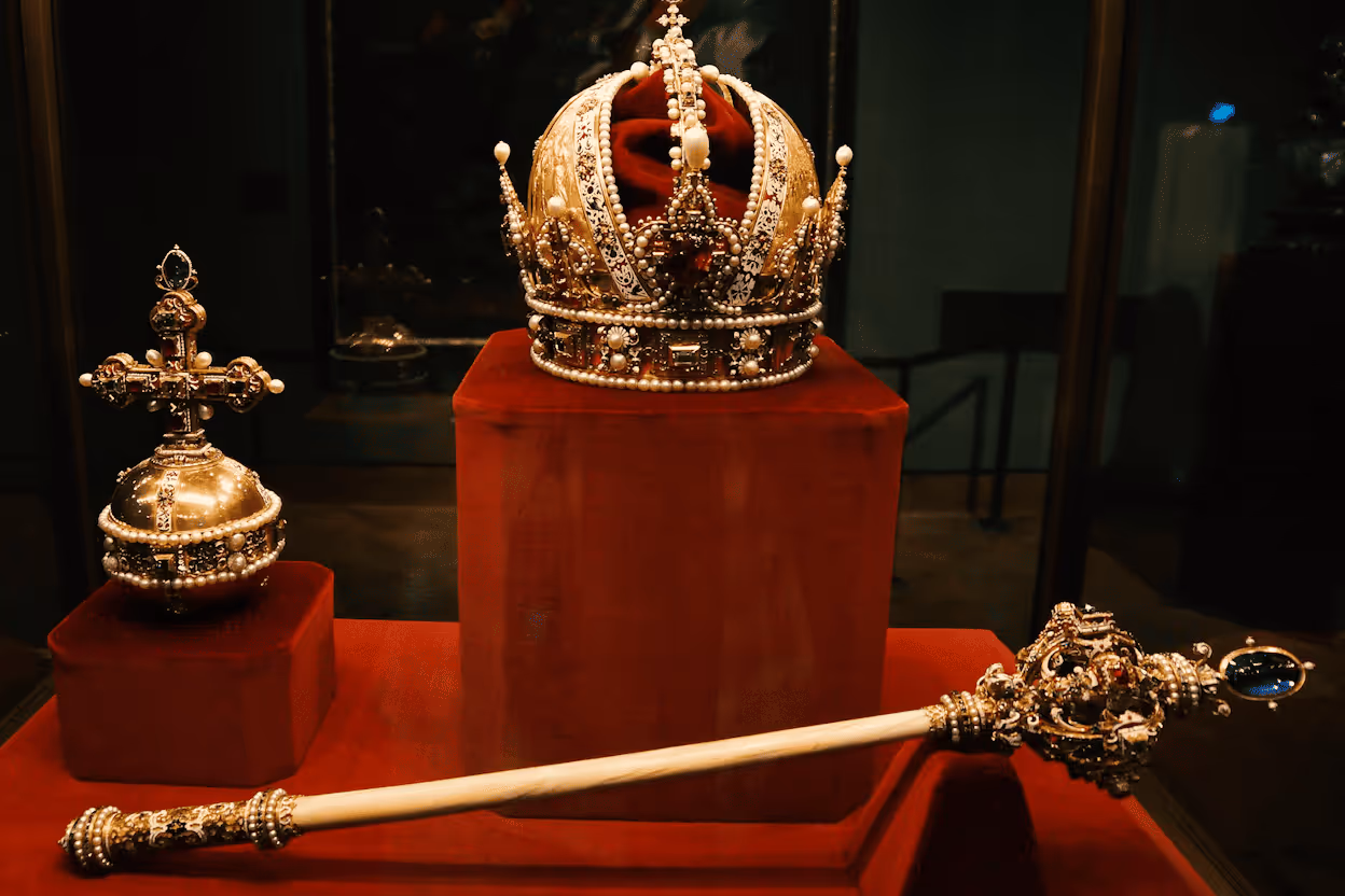 The Imperial Regalia of the Holy Roman Empire (or Austrian Empire) on display, featuring a central golden crown, an imperial orb on the left, and a scepter in the foreground, all heavily adorned with jewels and pearls on red velvet.