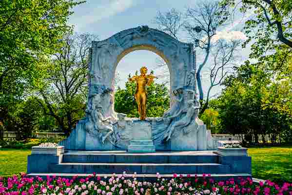 The Johann Strauss II Monument in Vienna's Stadtpark. A brilliant golden statue of the waltz composer playing his violin stands beneath a monumental white marble arch carved with dancing figures. The monument sits on a tiered pedestal, with a foreground of pink and white blooming tulips and a lush green park setting under a blue sky.
