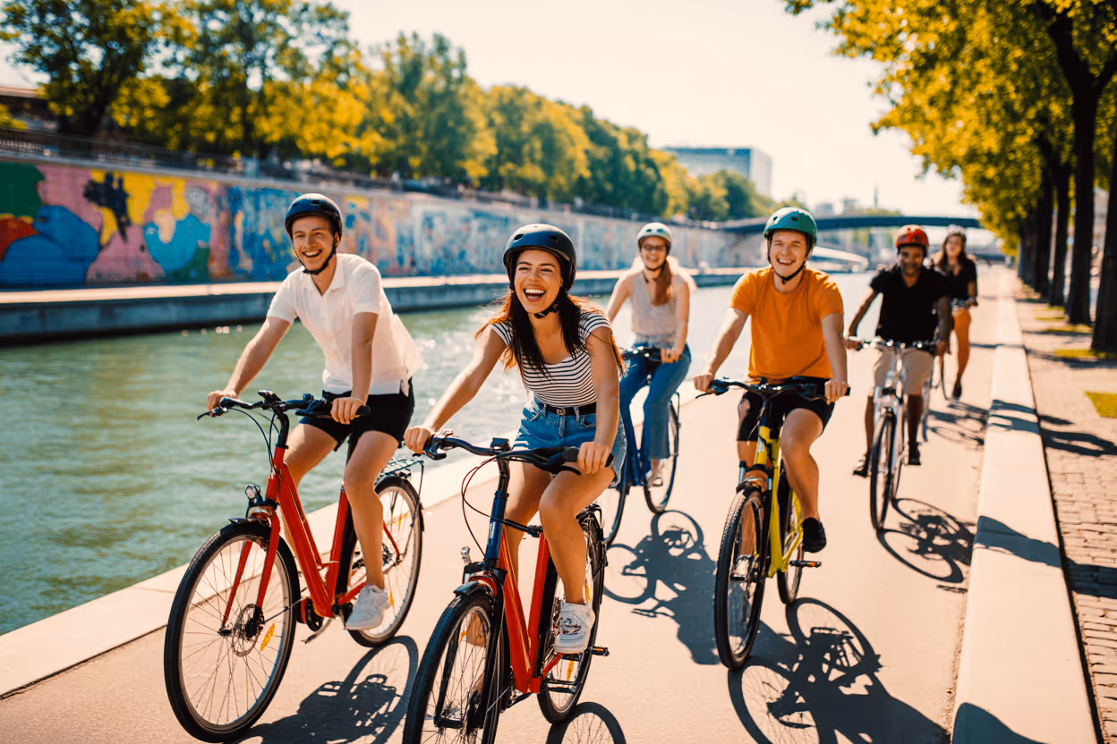 A sunlit photo of six friends riding bicycles along a waterfront path, possibly in a European city. The group, all wearing helmets, is smiling and laughing. The path is bordered by a canal or river on the left and a line of tall green trees on the right. Colorful graffiti is visible on the canal wall.