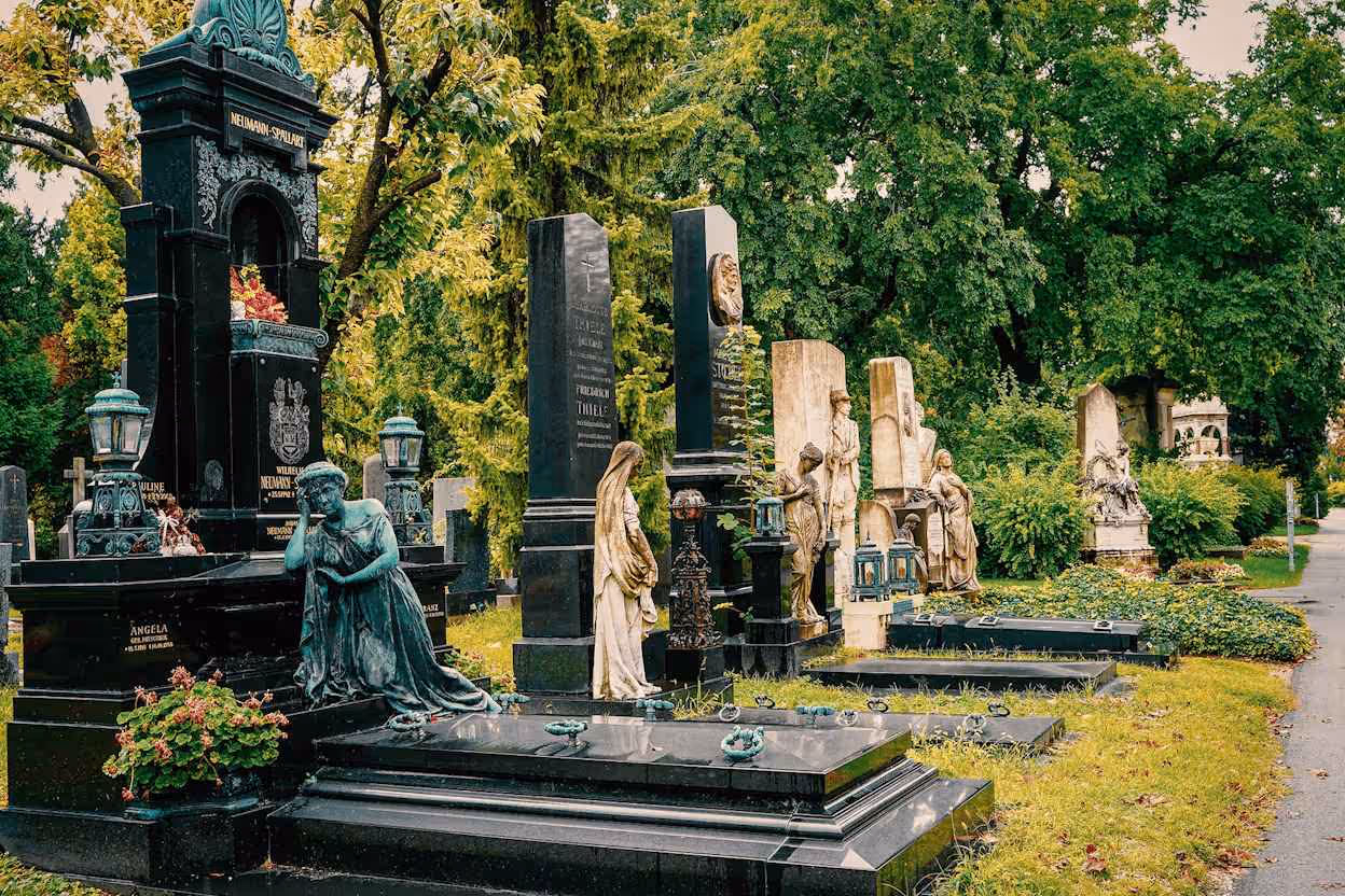 A view of a historic cemetery featuring rows of large, elaborate black marble monuments and statues, including a prominent kneeling female figure, set against a backdrop of lush green trees and lawn