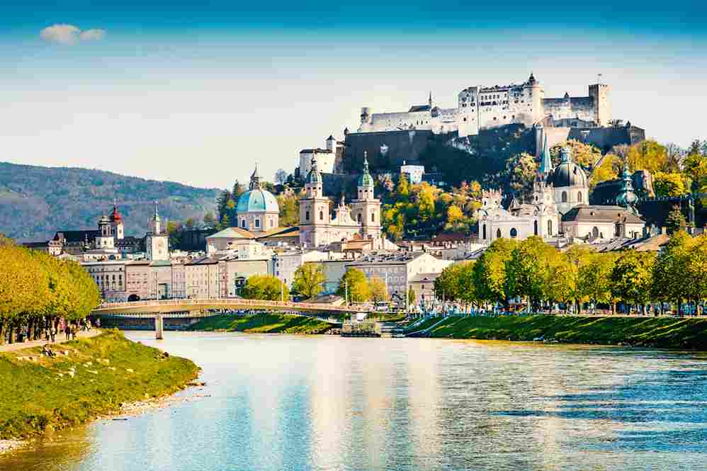 A panoramic view of Salzburg, Austria, showing the Salzach River, the historic city center with Baroque domes, and the large Hohensalzburg Fortress crowning the hill above.