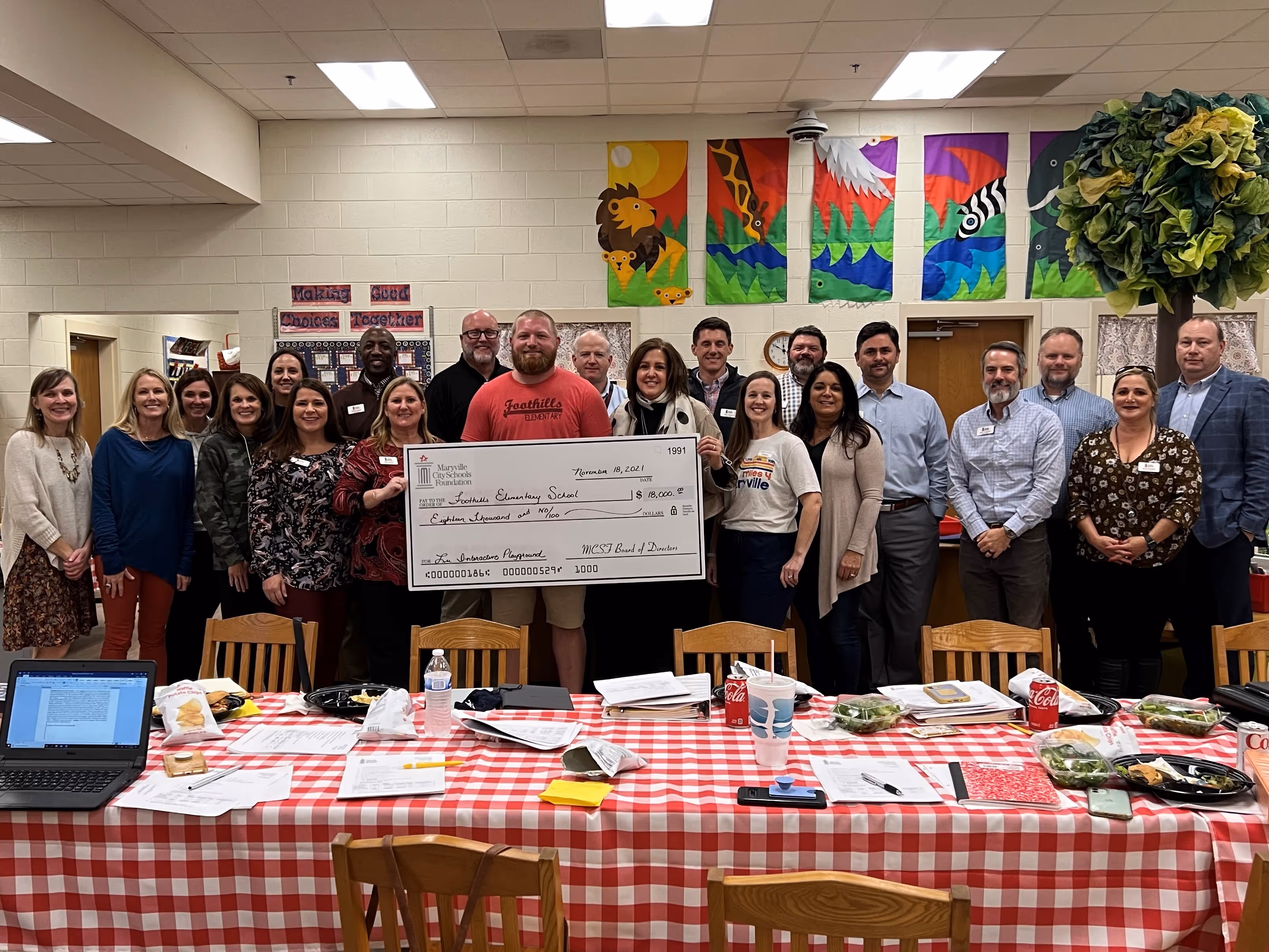 Group of adults standing behind a red and white checkered table, holding a large check for $18,000 made out to Foothills Elementary School for an interactive playground.