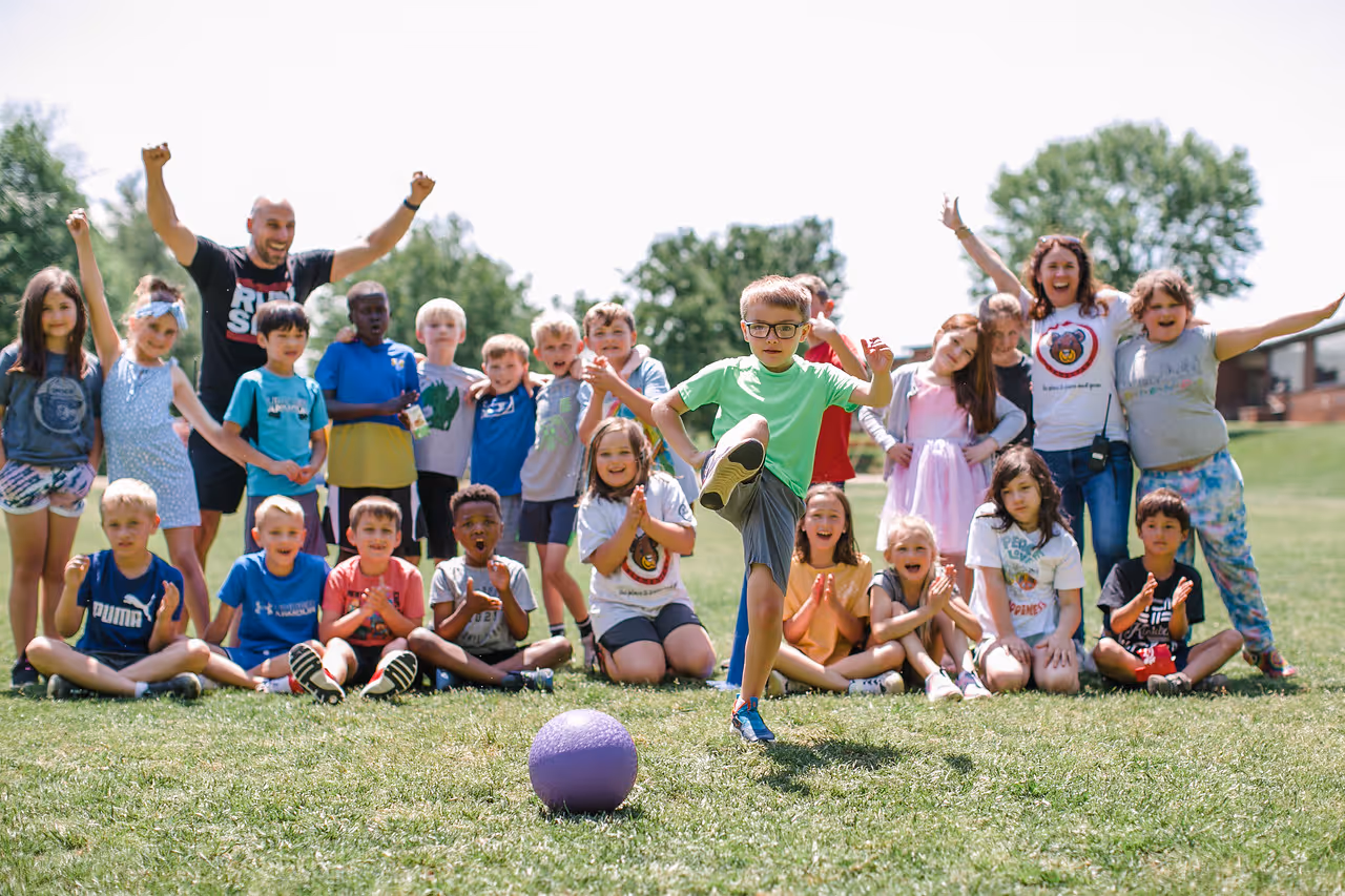A boy in a green shirt kicks a purple ball outdoors while a group of children and two adults cheer him on in the background.