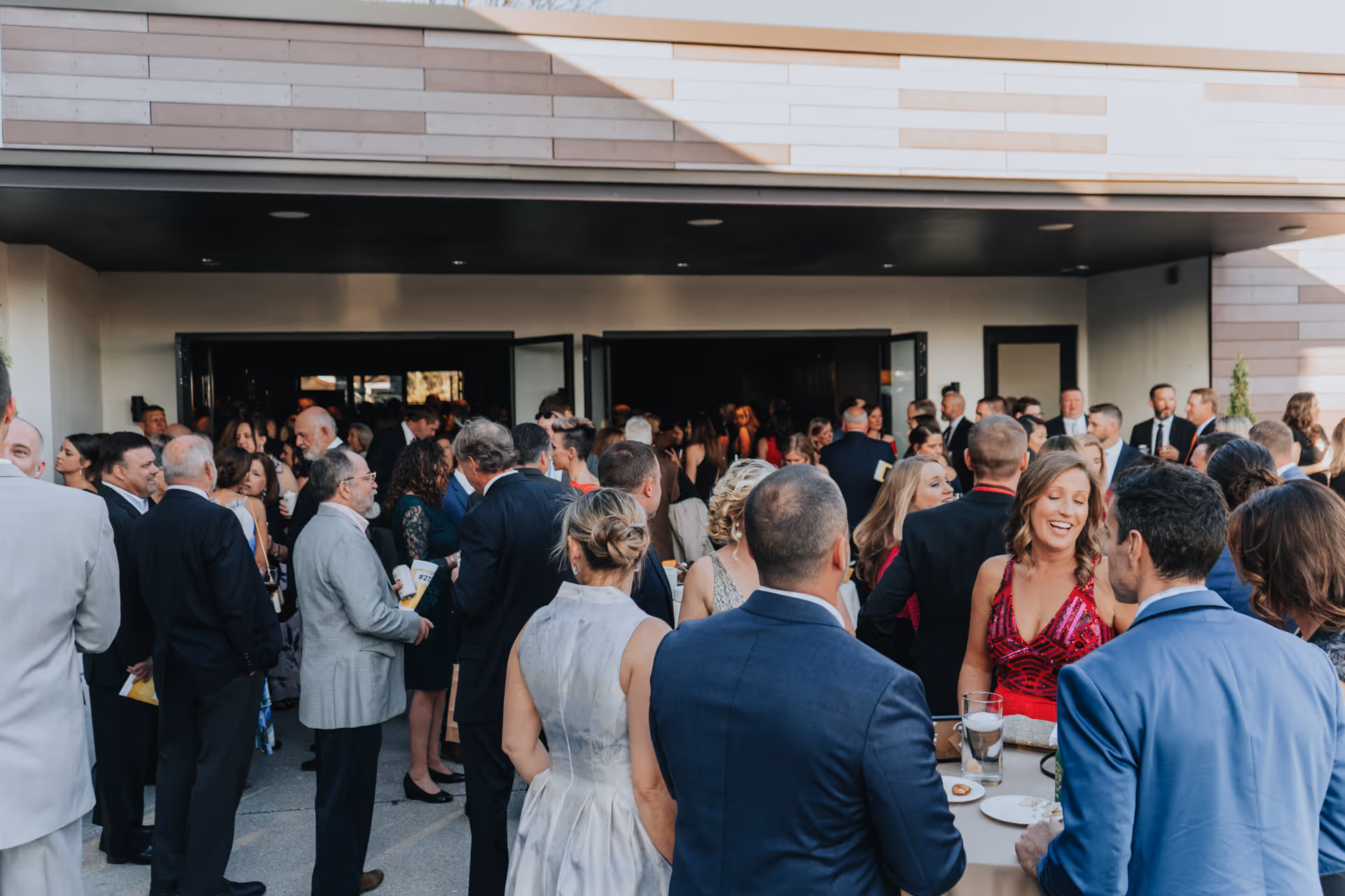 Crowd of people dressed in formal attire socializing outside a modern building with open doors.