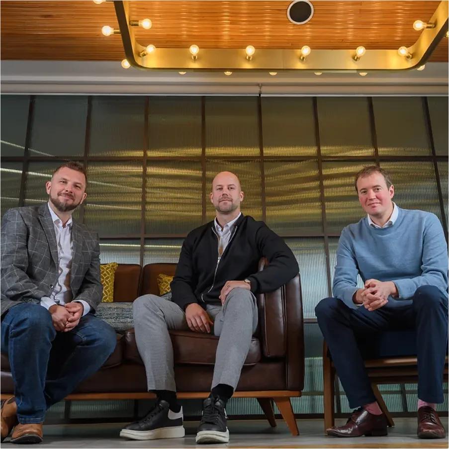 Three men sitting in a modern office space, with one on a brown leather sofa and two on chairs, under a wooden ceiling with lights.