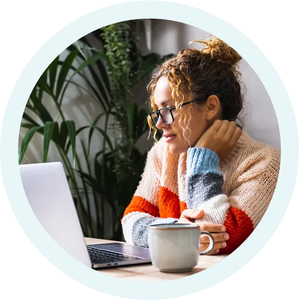 Woman with curly hair and glasses wearing a colorful knit sweater working on a laptop with a coffee mug in front of her.