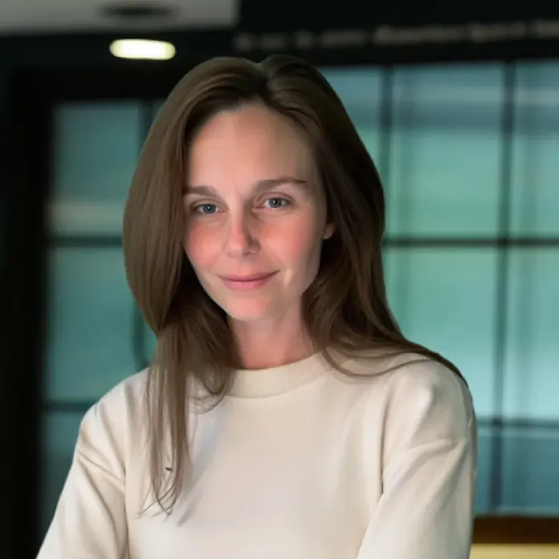 Woman with long brown hair wearing a cream sweater, smiling softly against a blurred indoor background with large windows.