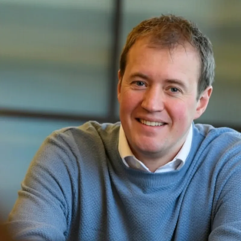 Smiling man with short light brown hair wearing a white shirt and blue sweater in an indoor setting.