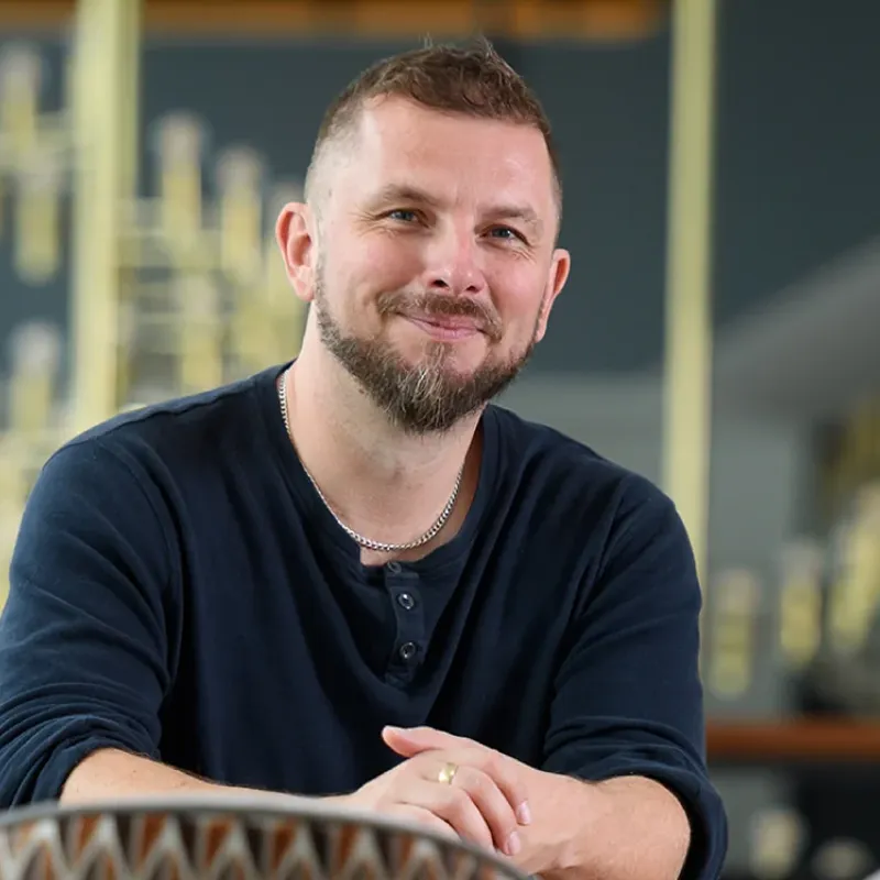 Smiling man with short hair and beard wearing a dark long-sleeve shirt and silver chain, sitting with hands clasped.