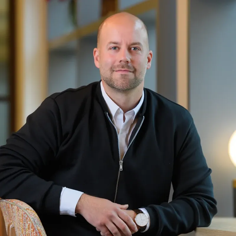 Smiling bald man in a black zip-up sweater over a white shirt sitting at a table with hands folded.