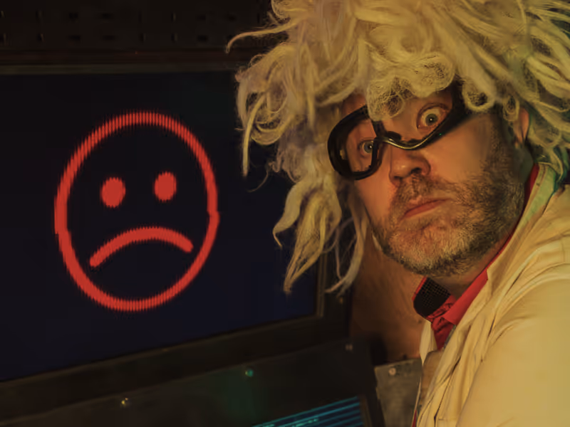 Scientist with wild white hair and goggles looks surprised beside a screen displaying a red sad face emoticon.