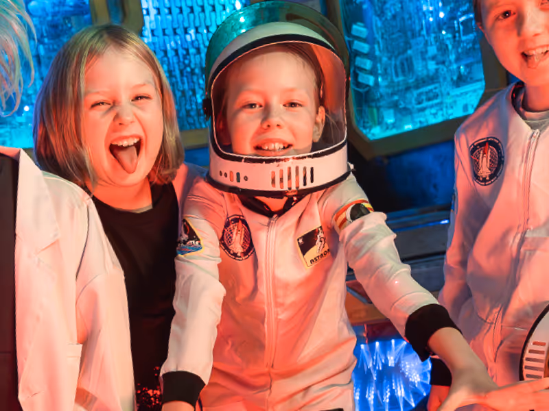 Three children smiling and wearing astronaut costumes in a space-themed setting.