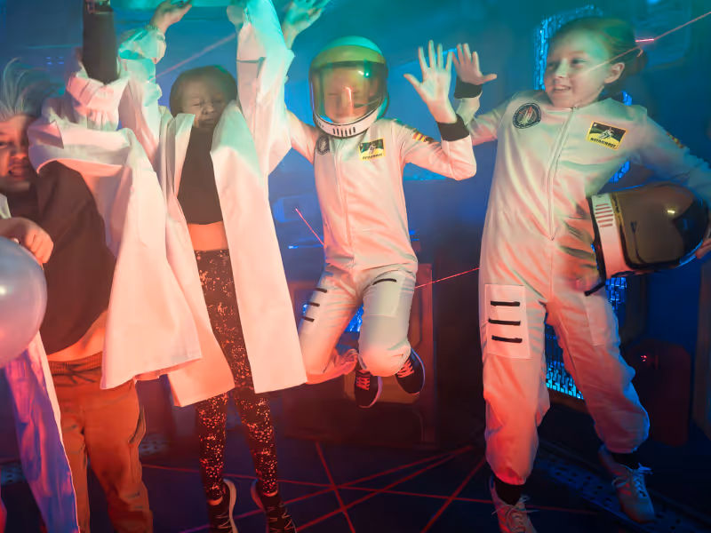 Four children dressed as astronauts and scientists jumping and playing in a dimly lit room with colored lights and laser beams.
