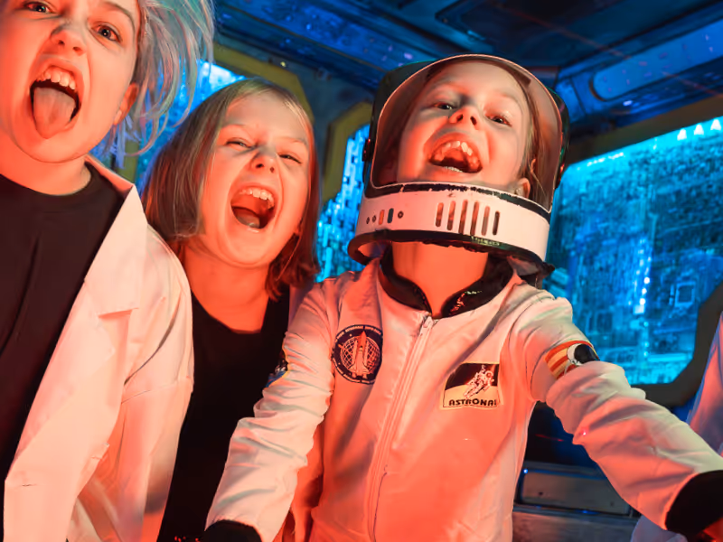 Three children laughing and making funny faces, one dressed as an astronaut inside a space-themed room.