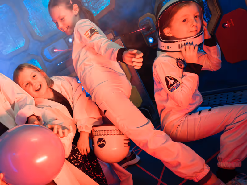 Three children happily dressed as astronauts inside a space-themed room with helmets and space suits.