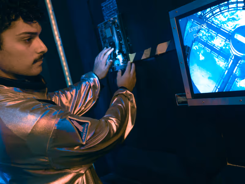A man in a metallic space suit working on a control panel with a monitor displaying a space station exterior view.