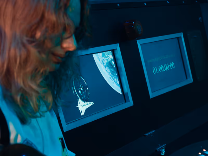 A player with curly hair wearing glasses looks at two screens showing Earth and a timer inside a dark control room.