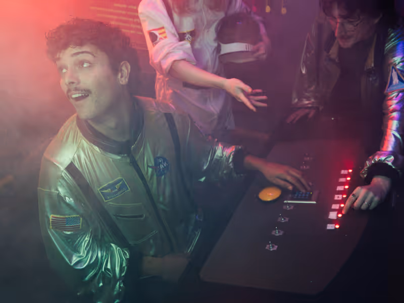 Three people in silver space-themed jackets interacting with futuristic control panels in a dimly lit room with red and purple lighting.